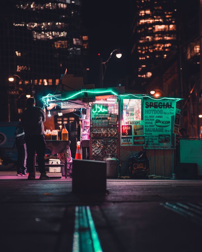 A vibrant street food stall illuminated by neon lights in a bustling city at night.